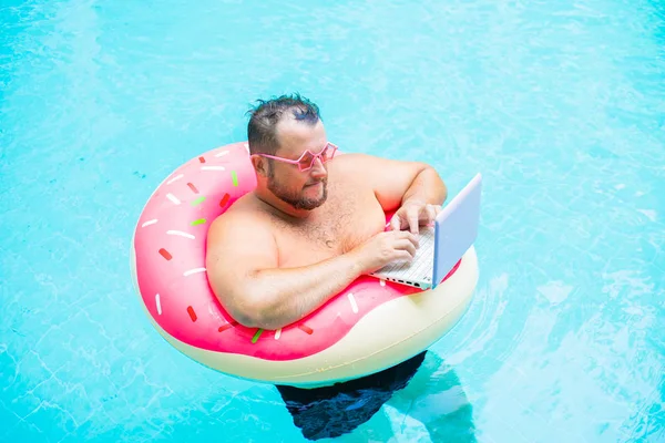 Ernstige grappige dikke man in roze bril op een opblaasbare cirkel in het zwembad werkt op een laptop portretend een meisje. — Stockfoto