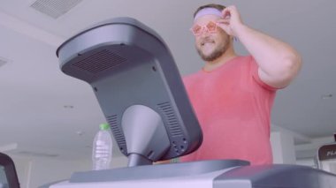 Pembe bir T-shirt ve pembe gözlük içinde oynak şişman adam spor salonunda fitness meşgul.