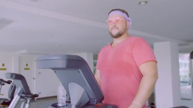 Pembe bir T-shirt ve pembe gözlük içinde oynak şişman adam spor salonunda fitness meşgul.