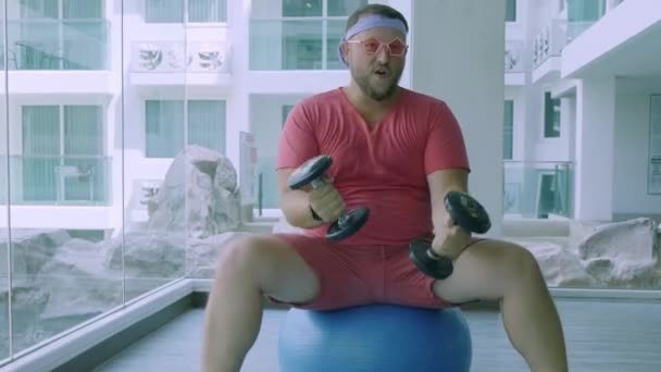 Playful fat man in a pink T-shirt and pink glasses is engaged in fitness with dumbbells and a fit ball in the gym. — Stock Video