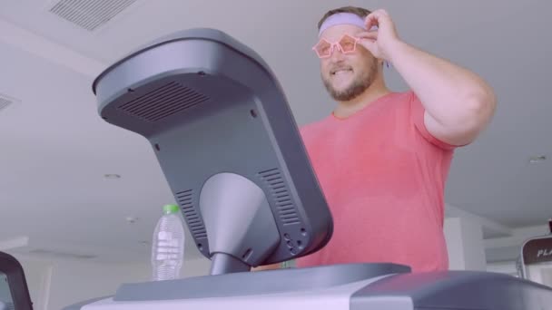 Hombre gordo juguetón en una camiseta rosa y gafas de color rosa se dedica a la aptitud en el gimnasio . — Vídeos de Stock