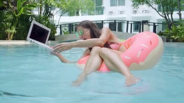 女人在游泳池的游泳圈里游泳，把笔记本电脑扔进水里。这个女孩正躺在一个装有微型电脑的充气戒指上。选择性聚焦 — 图库视频影像