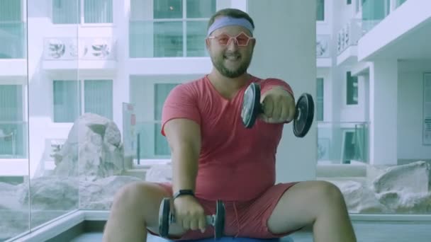 Homem gordo brincalhão em uma camiseta rosa e óculos rosa está envolvido na aptidão com halteres e uma bola de ajuste no ginásio . — Vídeo de Stock