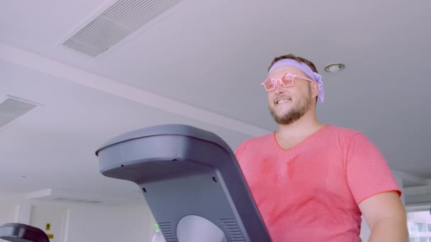 Speelse dikke man in een roze T-shirt en roze bril is bezig met fitness in de sportschool. — Stockvideo