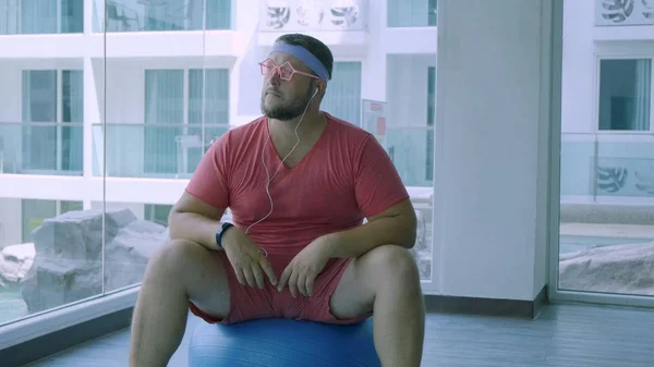 Playful fat man in a pink T-shirt and pink glasses is engaged in fitness with dumbbells and a fit ball in the gym. man listening to music on headphones.