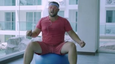 Pembe t-shirt ve pembe gözlük lütfedilen şişman adam halterve spor salonunda uygun bir top ile fitness yapan. kulaklıkla müzik dinleyen adam.