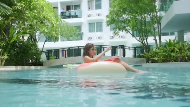 Женщина плавает в бассейне в бассейне и бросает ноутбук в воду. Девушка отдыхает на море на надувном кольце с мини-компьютером. селективный фокус — стоковое видео