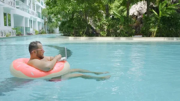 プールのインフレータブルサークルにピンクのメガネで深刻な面白い脂肪の男性は、女の子を描いたラップトップ上で動作します. — ストック写真