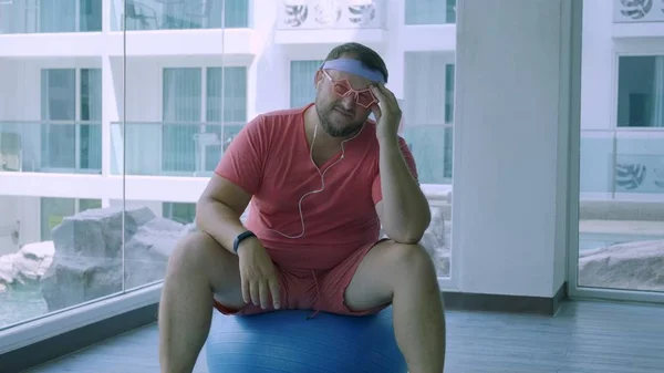 Playful fat man in a pink T-shirt and pink glasses is engaged in fitness with dumbbells and a fit ball in the gym. The man has a headache