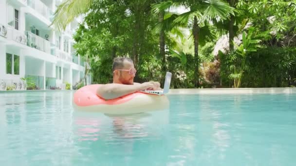 Grasso divertente uomo in cerchio gonfiabile rosa sorpreso e lascia cadere il computer portatile in acqua in piscina . — Video Stock