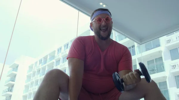 Playful fat man in a pink T-shirt and pink glasses is engaged in fitness with dumbbells and a fit ball in the gym.