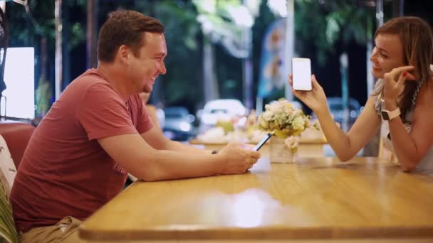 Couple in love man and woman are sitting in a cafe at the table on a date in the evening. Man shows woman something on the phone, girl smiles — Stock Video