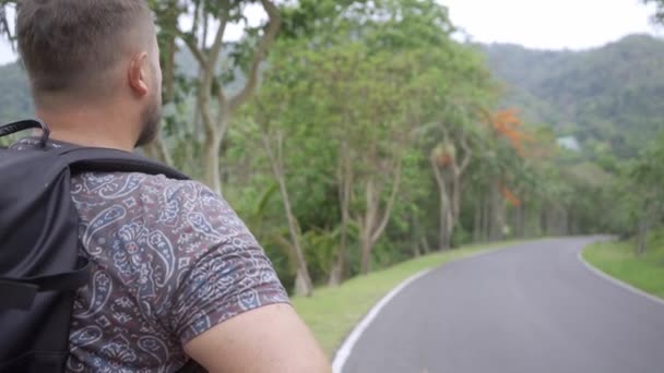 Bonita floresta tropical com um jovem viajante com uma mochila na estrada para a floresta da Tailândia. Um homem caminha em uma estrada tropical e desfruta das vistas da natureza — Vídeo de Stock