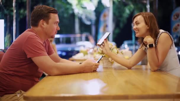 Para w miłości mężczyzny i kobiety siedzą w kawiarni przy stole na randkę wieczorem. Mężczyzna pokazuje kobietę coś na telefon, Dziewczyna uśmiecha się — Wideo stockowe