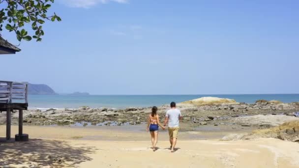 Una pareja amorosa camina a lo largo de la arena dorada de la playa, caminan sobre piedras, se ayudan mutuamente y se toman selfies. Vista desde arriba — Vídeo de stock