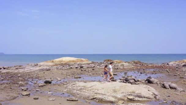 Een liefdevol paar loopt langs het gouden zand van het strand, lopen op stenen, helpen elkaar en nemen selfies. Uitzicht vanaf boven — Stockvideo