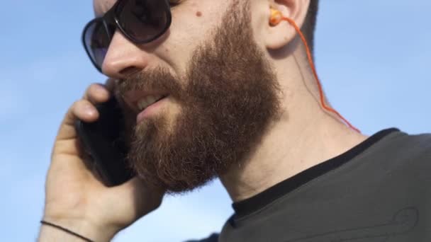 Homme courageux avec une barbe dans les lunettes, parle nerveusement au téléphone contre le ciel bleu dans l'air frais. Gros plan . — Video