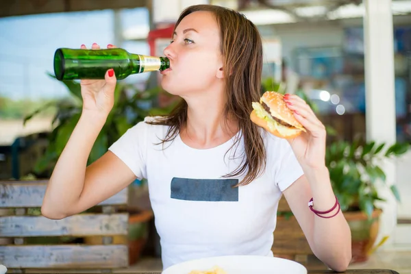 Young attractive girl, with an independent look, drinks beer and eats a burger. Close up. The concept of feminism and womens independence. — Stock Photo, Image