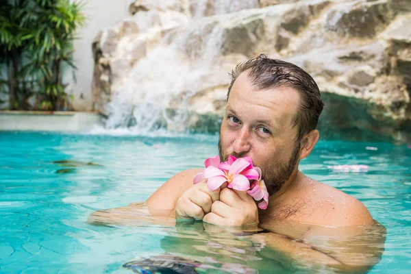 Mavi su ile bir havuzda sakallı Charming adam, elinde bir plumeria çiçek tutar ve gülümsüyor.