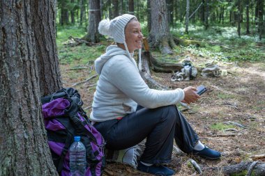 Hafif giysiler içinde güzel bir kız, Avrupalı görünümlü bir ekoturist, ormanda bir ağacın yanında oturur ve telefonbakar.
