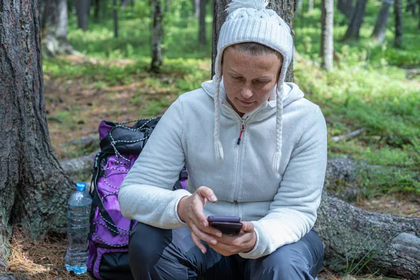 Hafif giysiler içinde güzel bir kız, Avrupalı görünümlü bir ekoturist, ormanda bir ağacın yanında oturur ve telefonbakar.