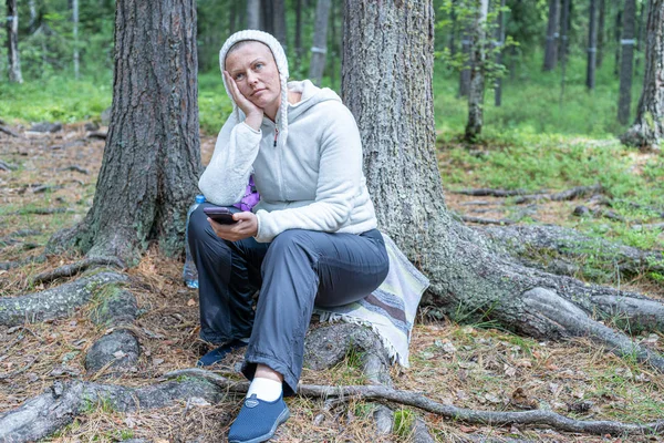 Hafif giysiler içinde güzel bir kız, Avrupalı görünümlü bir ekoturist, ormanda bir ağacın yanında oturur ve telefonbakar.