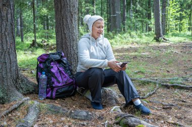 Hafif giysiler içinde güzel bir kız, Avrupalı görünümlü bir ekoturist, ormanda bir ağacın yanında oturur ve telefonbakar.