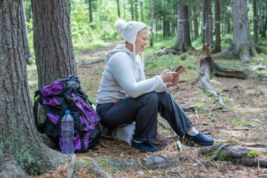 Hafif giysiler içinde güzel bir kız, Avrupalı görünümlü bir ekoturist, ormanda bir ağacın yanında oturur ve telefonbakar.