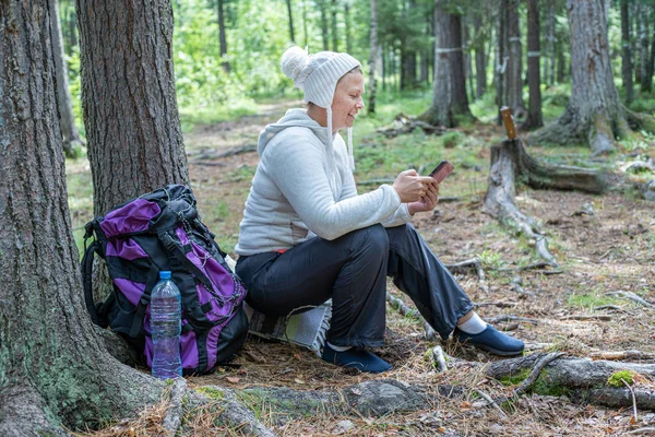 Hafif giysiler içinde güzel bir kız, Avrupalı görünümlü bir ekoturist, ormanda bir ağacın yanında oturur ve telefonbakar.
