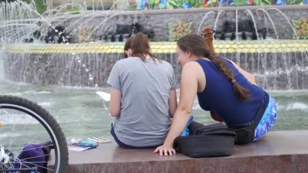 Due ragazze LGBT vicino alla fontana, vista posteriore, una con la chitarra, l'altra dipinge . — Video Stock