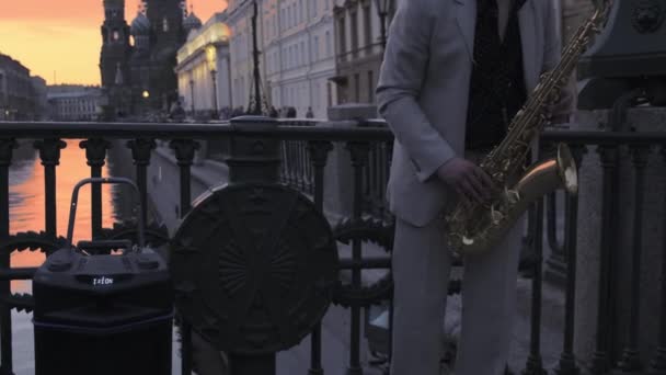 Saxofonista em um terno leve em uma ponte no pôr-do-sol, toca o saxofone . — Vídeo de Stock