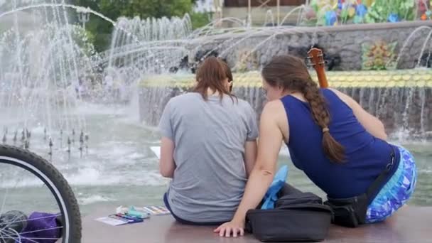 Dos chicas LGBT cerca de la fuente, vista trasera, una con una guitarra, las otras pinturas . — Vídeo de stock