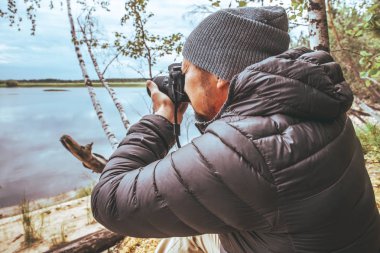 Gri ceketli bir adam, nehrin kıyısında, ormanda doğayı fotoğraflar.