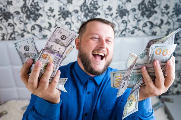 A nice man with a beard in a blue shirt, rejoices at the dollars, throws them up and counts. Win lottery — Stock Photo, Image