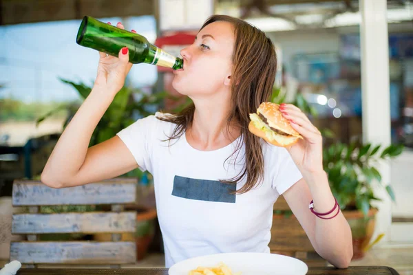 Jong aantrekkelijk meisje, met een onafhankelijke blik, drinkt bier en eet een hamburger. Close-up. Het concept van feminisme en Womens onafhankelijkheid. — Stockfoto