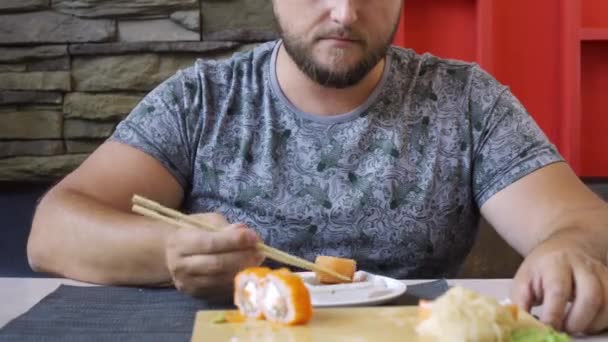Homem gordinho agradável com barba em um bar de sushi, rolos de comer engraçados. Não sabe como lidar com paus, rolos caem . — Vídeo de Stock
