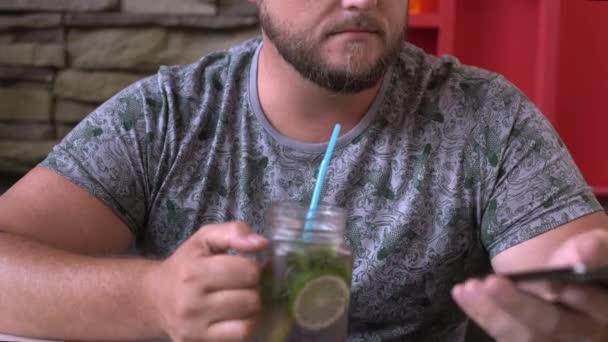 Приємний пухкий чоловік Європейського вигляду, випиваючи коктейль в кафе, дивлячись на телефони посміхаючись. — стокове відео
