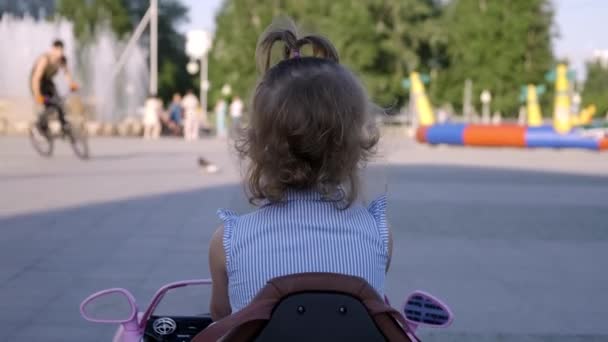 Niña pequeña en un vestido azul conduciendo un coche de juguete rosa en el parque y sonriendo, de cerca — Vídeo de stock