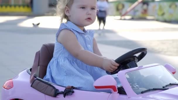 Bambina con un vestito blu che guida una macchinina rosa nel parco e sorridente, da vicino — Video Stock