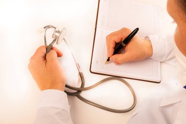 Doktor beyaz bir masada bir ofiste oturur, bir not defterinotlar yapar, bir stetoskop yakın, yukarıdan görünümü yatıyor. Yakın çekim