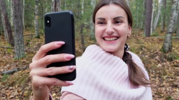 Una joven mujer feliz toma selfie en el teléfono con placer en el bosque de primavera y sonríe. Más despacio. De cerca. — Vídeo de stock