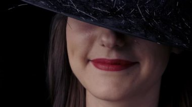 Siyah cadı şapkalı, parlak kırmızı dudakları gülümseyen çekici gizemli bir kadının yarım taraflı yüzünün yakın çekim. Cadılar Bayramı konsepti.