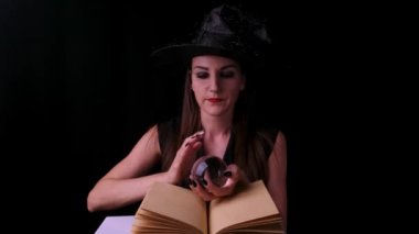 Siyah arka planda parlak kırmızı dudaklı çekici mistik cadı bir büyü kitabının yanında büyü yaparken sihirli bir topa bakıyor.