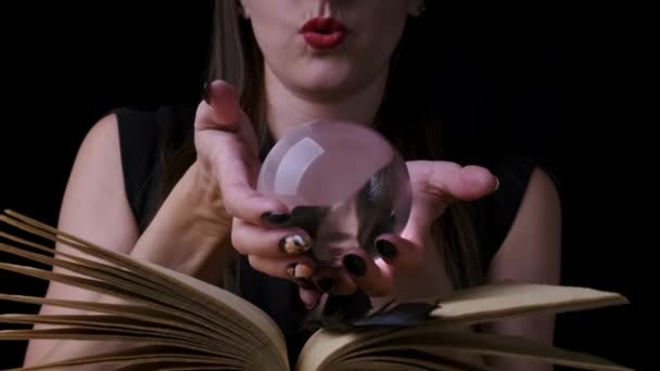Mulher bruxa mística atraente com lábios vermelhos brilhantes em um fundo preto, olha para uma bola mágica ao lançar feitiços ao lado de um livro de feitiços — Vídeo de Stock
