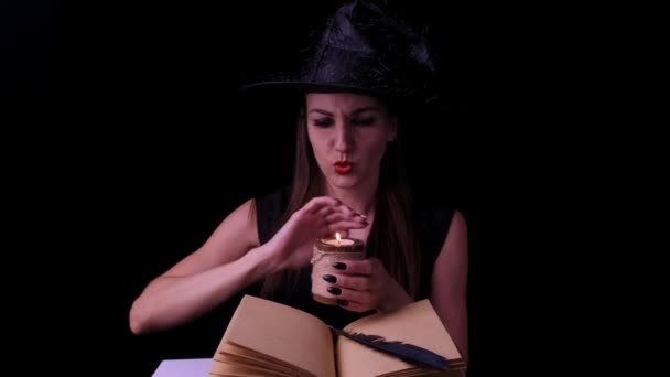 Attractive witch, on a black background, holding a candle in her hands over a spell book — Stock Video