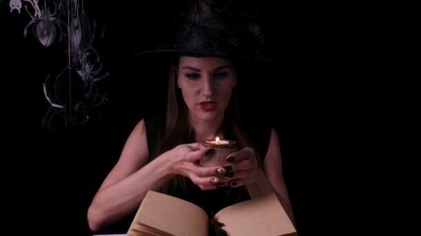 Привлекательная мистическая колдунья с ярко-красными губами на черном фоне, смотрит в волшебный шар во время заклинаний рядом с книгой заклинаний — стоковое видео