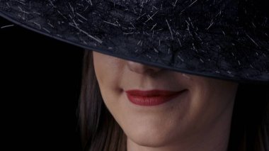 Siyah cadı şapkalı, parlak kırmızı dudakları gülümseyen çekici gizemli bir kadının yarım taraflı yüzünün yakın çekim. Cadılar Bayramı konsepti.