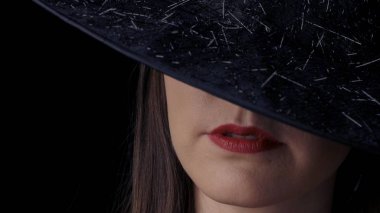 Siyah cadı şapkalı, parlak kırmızı dudakları gülümseyen çekici gizemli bir kadının yarım taraflı yüzünün yakın çekim. Cadılar Bayramı konsepti.