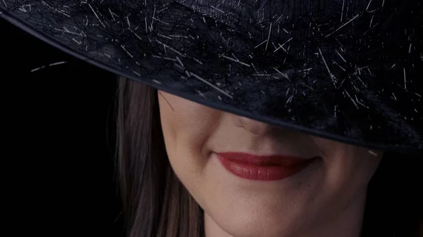 Blízký obličej atraktivní a záhadné ženy v černém čarodějném klobouku s zářivě červenými rty usmívajícími se. Halloweenský koncept. — Stock fotografie