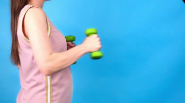 Bir kadın mavi arka planda yeşil halterlerle egzersiz yapıyor.. 